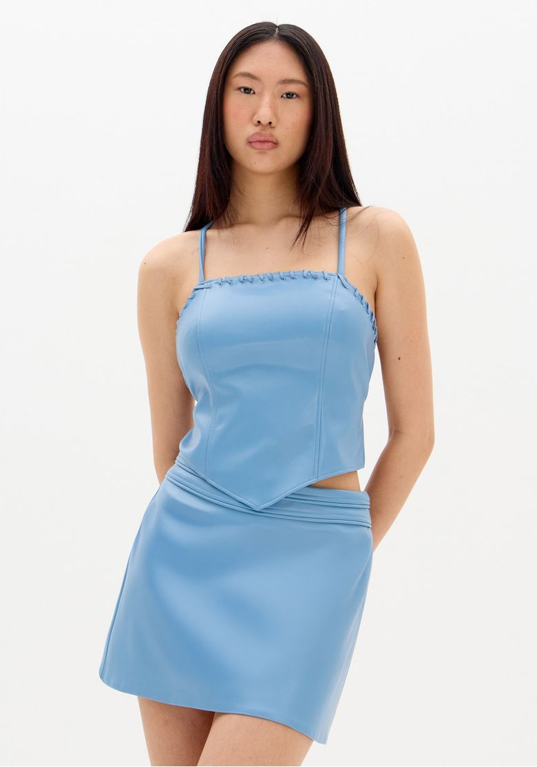 Modelo frontal mostrando blusa cropped azul em PU com alças finas, barra em bico e saia combinando, visual completo em estúdio branco.