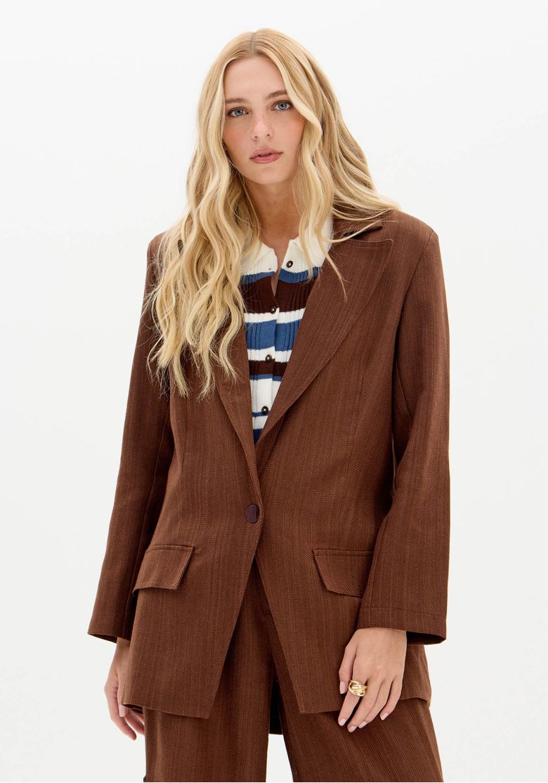 Modelo usando blazer marrom alongado frente com lapela clássica e bolsos com aba, look trabalho em tecido plano pesado