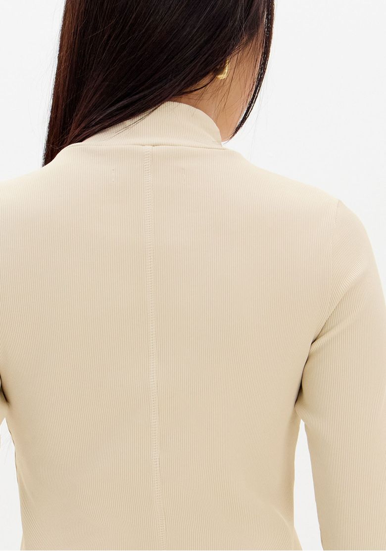 Close no decote alto da blusa gola alta canelada bege com bordado discreto aplicado no peito e textura canelada visível