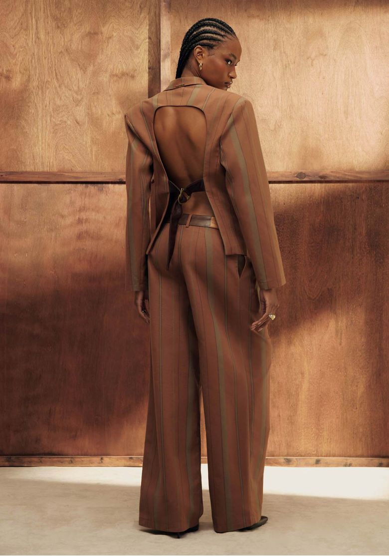 Modelo vista traseira com blazer oversized listrado marrom, destaque para a abertura nas costas e calça pantalona, cenário com painel de madeira