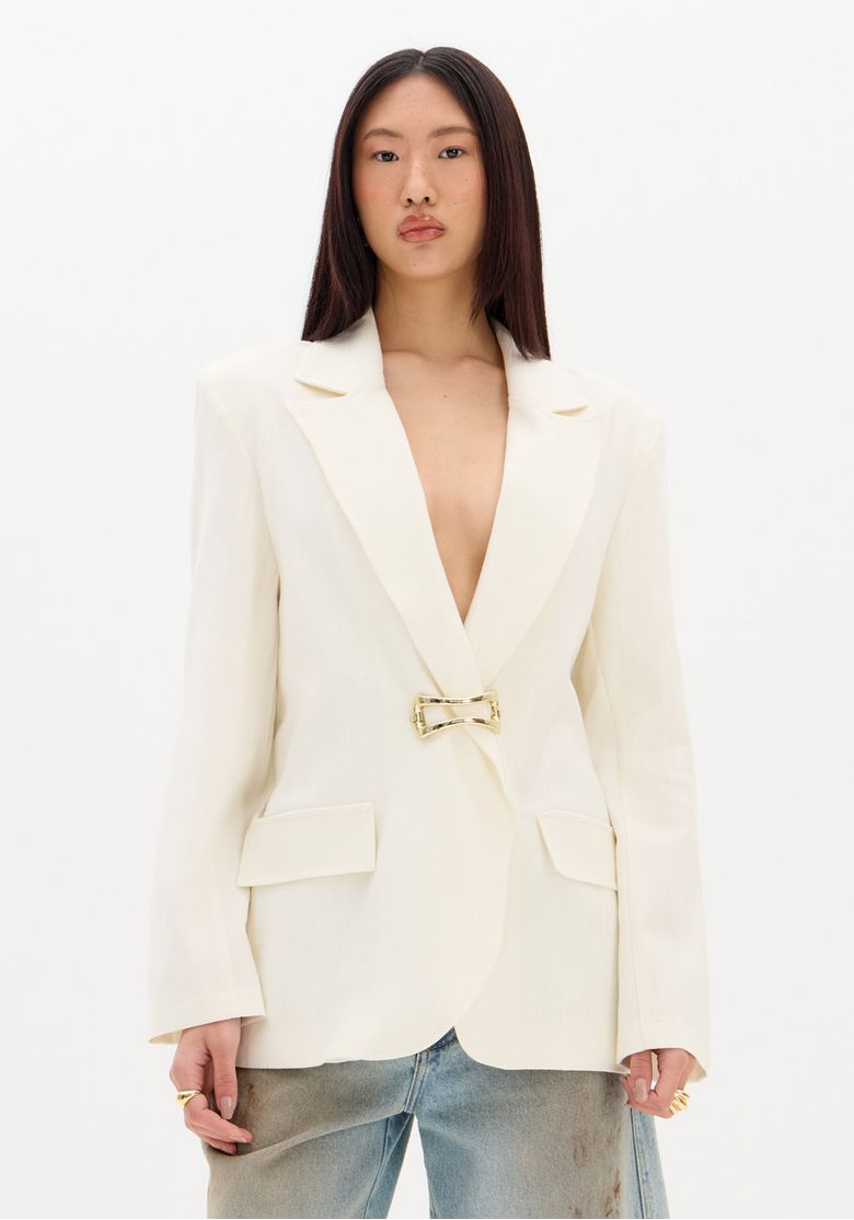 Modelo vestindo blazer branco alongado com fechamento metálico e bolsos com aba, vista frontal.