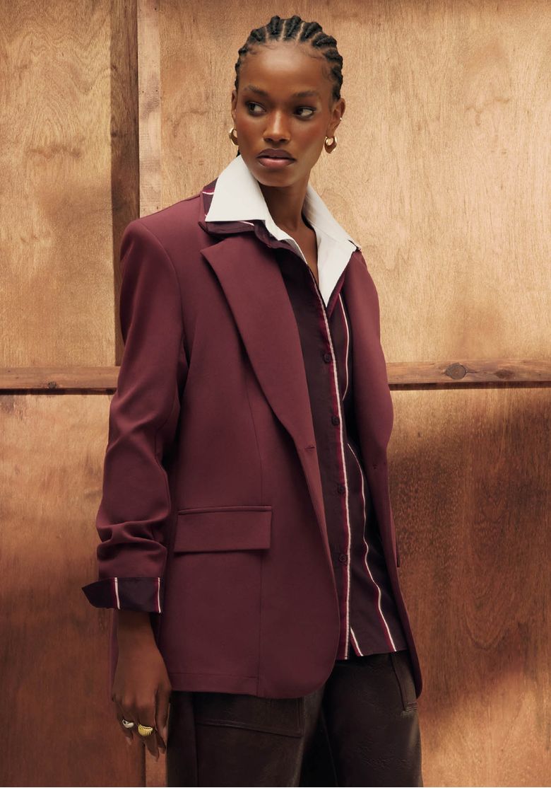 Modelo negra em três quartos usando blazer super oversized vermelho alongado com ombreiras, lapela clássica e bolsos com aba em fundo de madeira