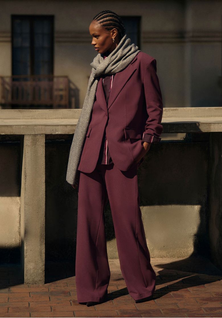 Modelo loira em estúdio usando blazer oversized vermelho alongado com calça listrada vertical vinho e branco e grande cachecol cinza drapeado no ombro