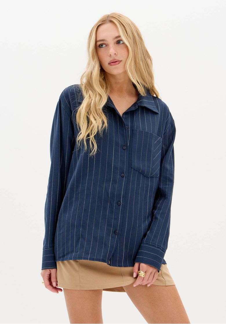 visao-frontal-da-camisa-oversized-azul-risca-de-giz-parcialmente-para-dentro-de-saia-bege-mostrando-gola-tradicional-e-abotoamento-discreto