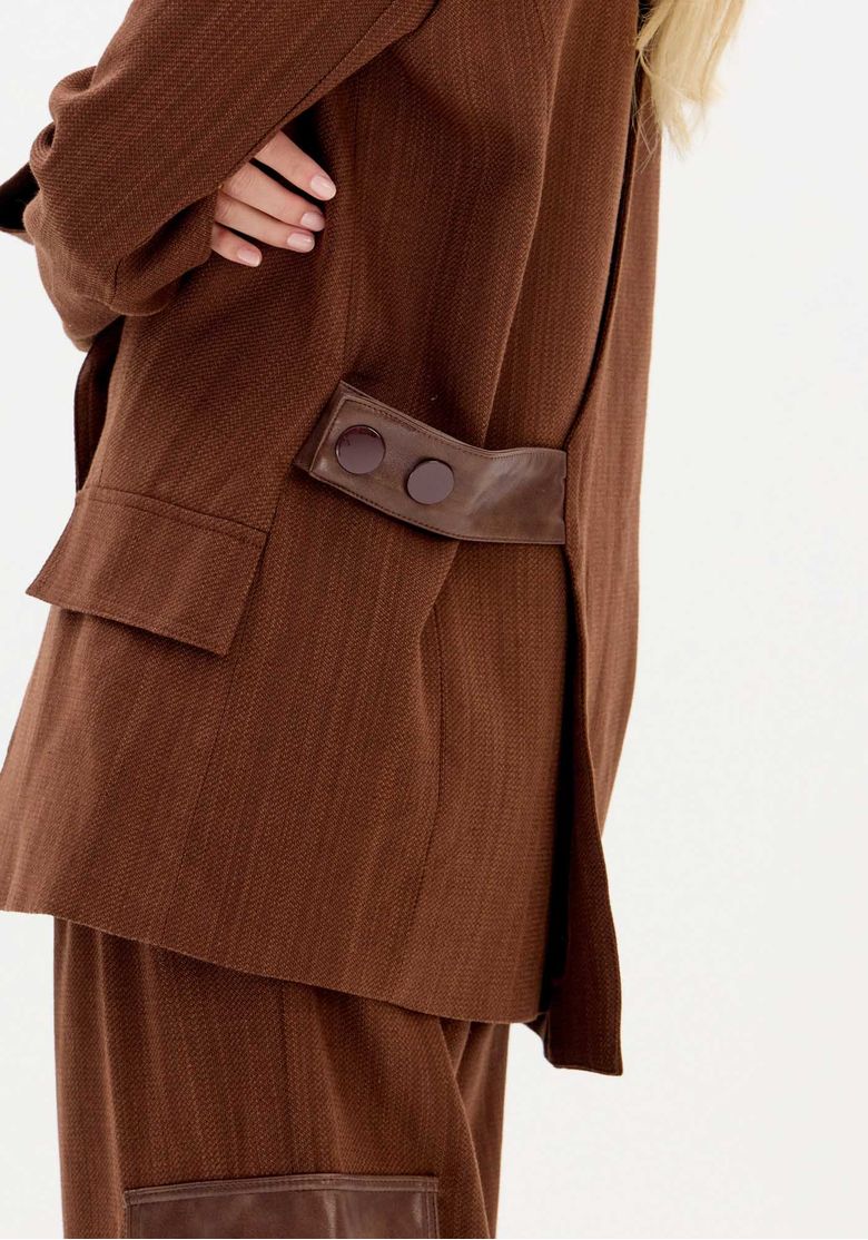 Close do detalhe de tira em couro com botões grandes e bolso com aba no corpo do blazer marrom