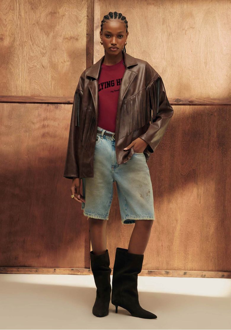 Modelo vestindo casaco oversized marrom com franjas, combinado com bermuda jeans e botas pretas.