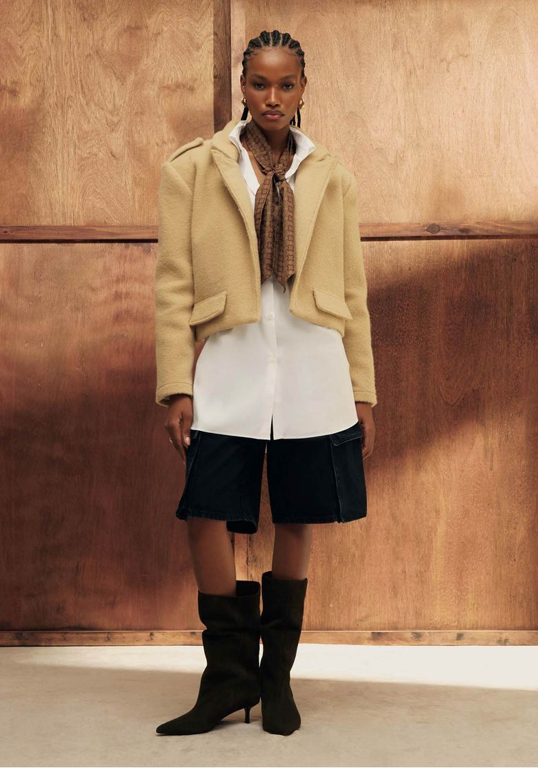 Modelo negra trajando casaco cropped oversized bege sobre camisa longa branca e bermuda preta, compondo um estilo urbano em frente a um fundo de madeira.