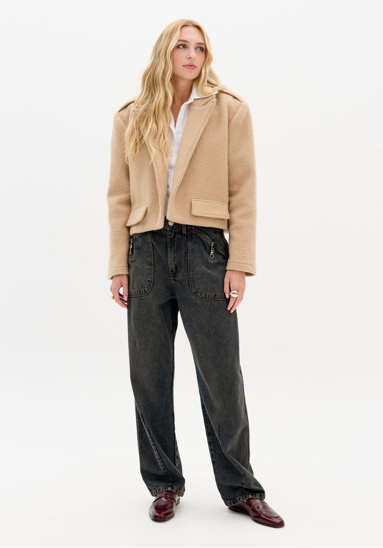 Modelo loira vestindo casaco cropped oversized bege, mostrando detalhe da gola de lapela e bolsos com aba visíveis.