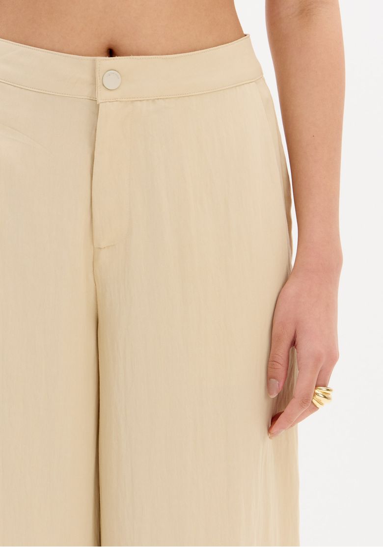 Detalhe frontal da calça slouchy bege mostrando pregas sutis, cós alto com fechamento por botão e caimento amplo