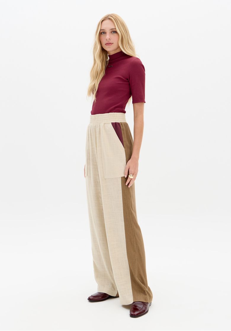 Modelo em pé, vista lateral usando calça reta em linho bicolor com cós elástico e bolsos frontais sobrepostos, recortes verticais marrom e bege