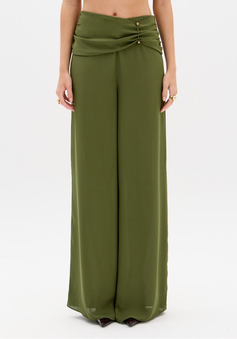Close da pala drapeada na cintura média da calça pantalona verde, com pregas, botões dourados em destaque e acabamento impecável no cós.