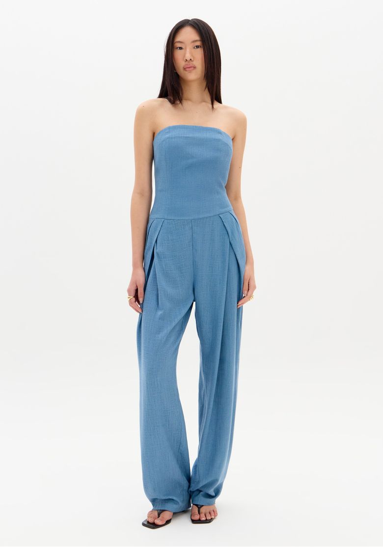 Modelo em pé usando macacão longo sem alça azul, vista frontal inteira com pernas pantalona e bolsos laterais, tecido linho leve