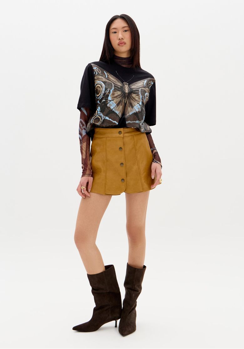 Modelo-em-pe-vestindo-short-saia-PU-bege-com-botoes-frontais-e-camiseta-preta-estampada-visual-casual-urbano-acompanhado-por-botas-marrom-escuras.