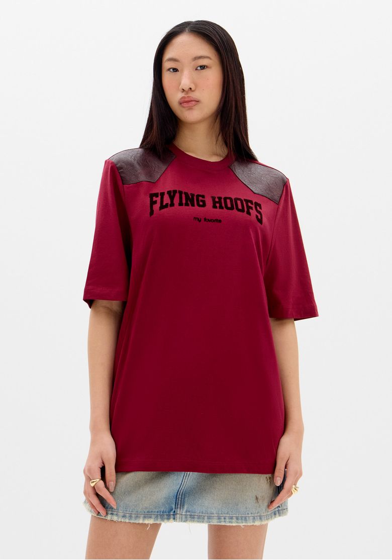 Camiseta oversized bicolor vermelha vista frontal em modelo, recortes contrastantes nos ombros em poliuretano e estampa 'FLYING HOOFS' em destaque