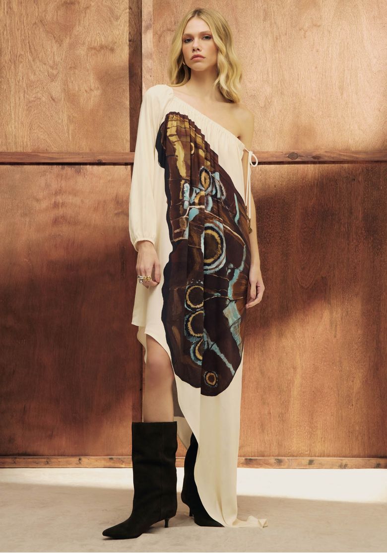 Modelo vestindo vestido curto assimétrico com estampa exclusiva em fundo bege, detalhe de ombro único.