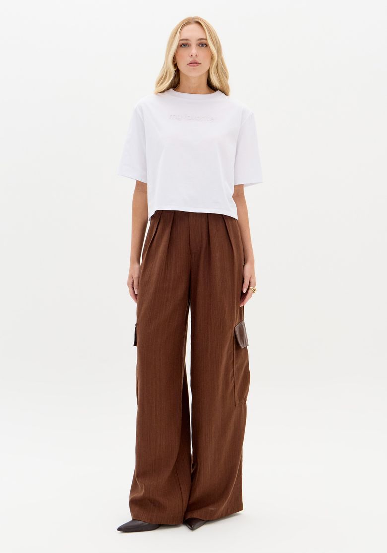 Look com camiseta oversized branca combinada com calça pantalona marrom, vista de corpo inteiro em cenário minimalista