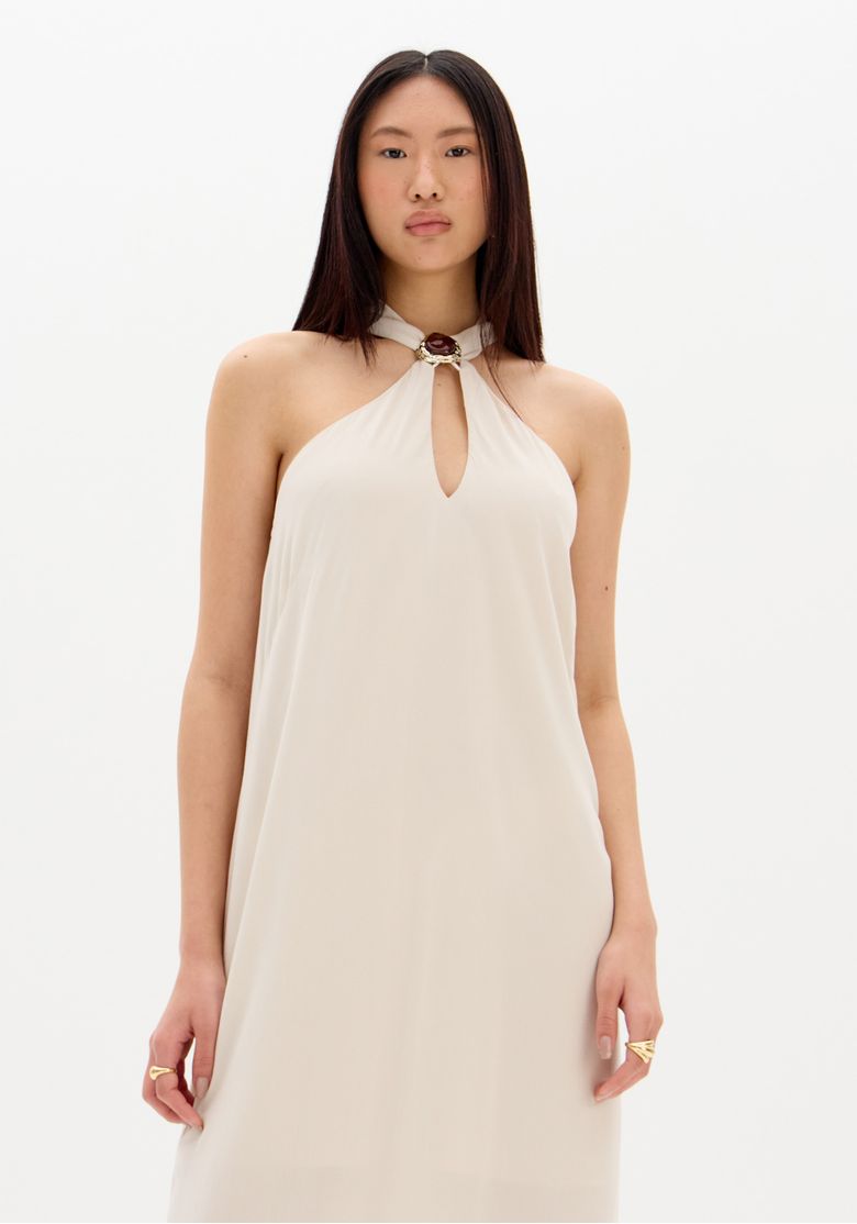 Close do detalhe do decote do vestido super midi bege com aplicação metálica decorativa no pescoço e tecido de acabamento suave