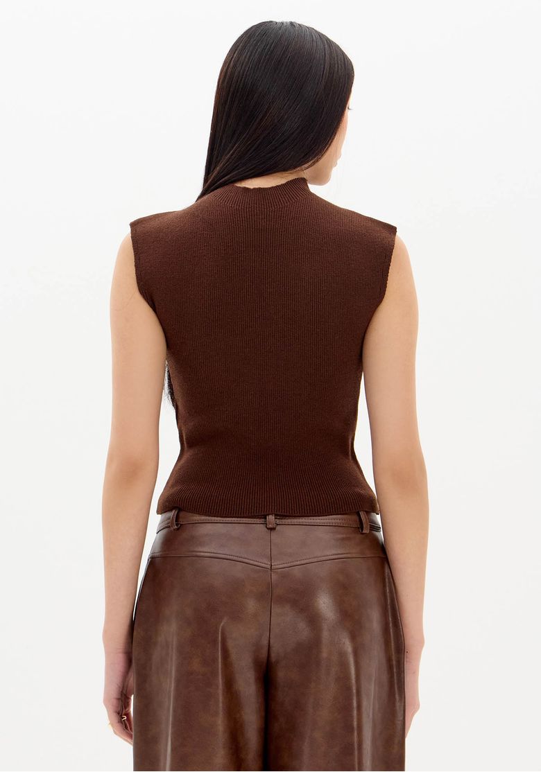 Modelo em pé usando blusa tricô sem mangas marrom de gola alta combinada com calça pantalona de couro marrom, look casual urbano em estúdio