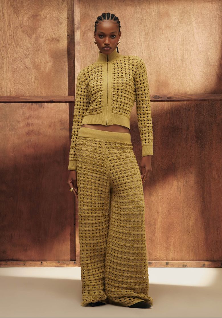 Modelo com conjunto de tricô bege em estúdio com fundo de madeira, jaqueta cropped e calça reta em ponto vazado - look completo