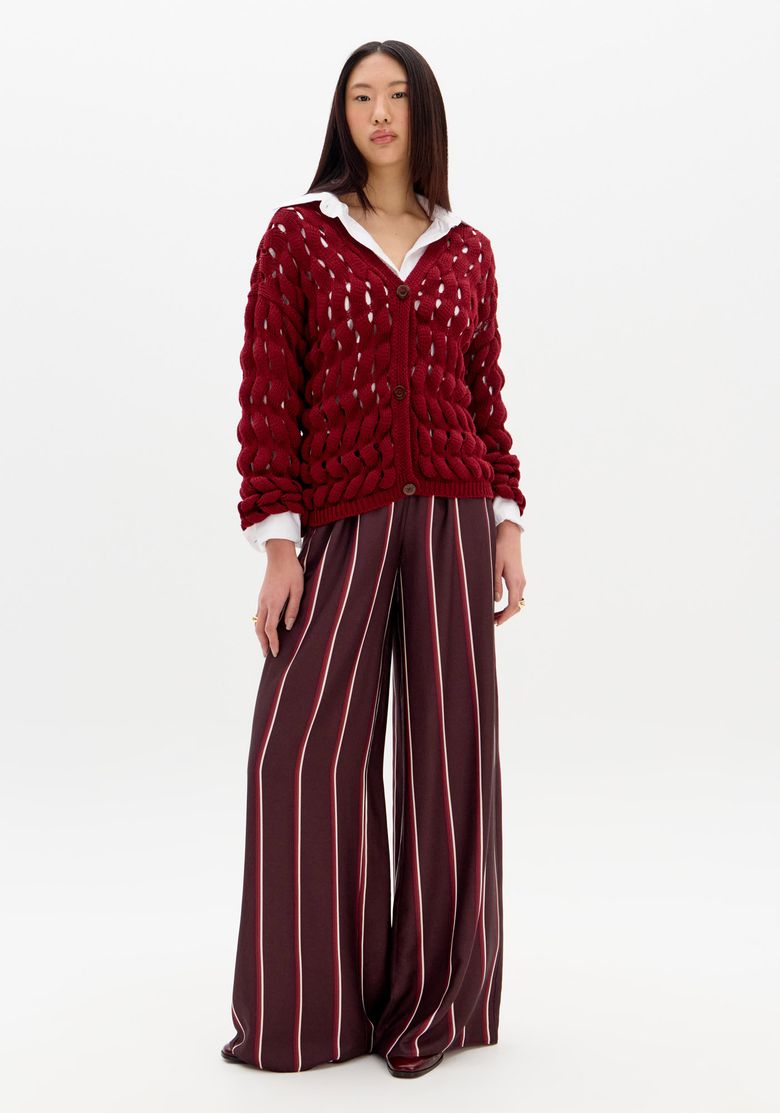 Modelo vestindo cardigã vermelho em tricô vazado com calça listrada, mostrando o visual completo.