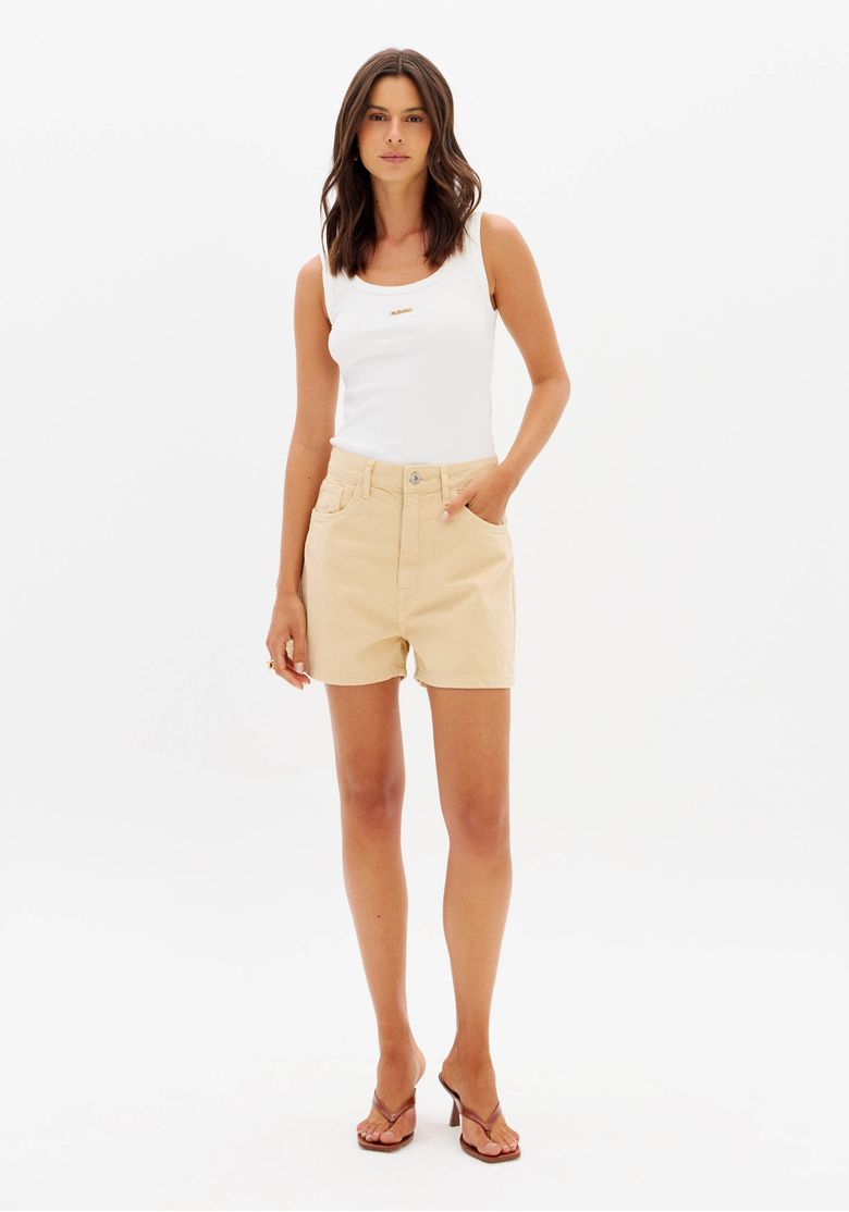 Modelo em estúdio usando short sarja mom bege com regata branca, vista frontal mostrando cintura super alta, comprimento curto e bolsos funcionais
