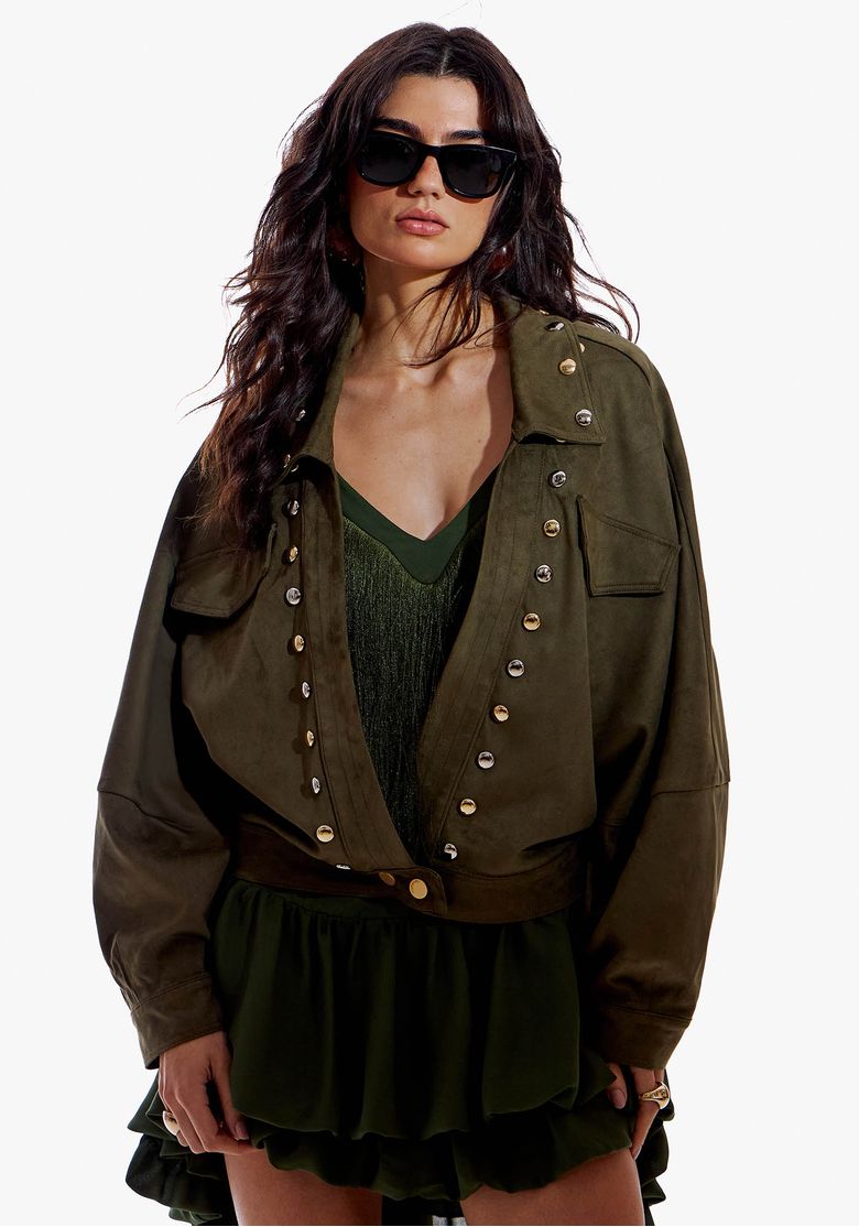 Modelo usando jaqueta de suede verde escuro com tachas metálicas na frente, gola de lapela e bolsos com lapela, look casual urbano.