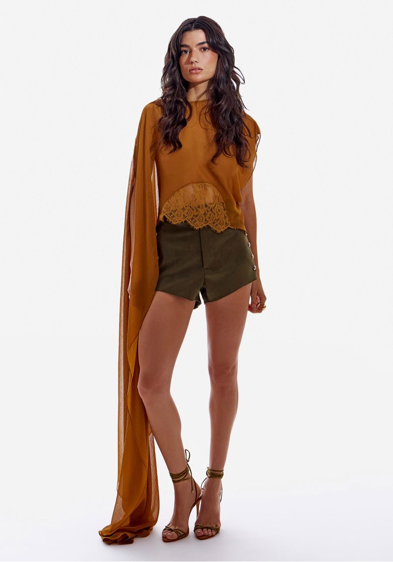 Modelo em pé vestindo short hot pants de suede verde escuro com blusa caramelo e sandálias de tiras, visual frontal completo.