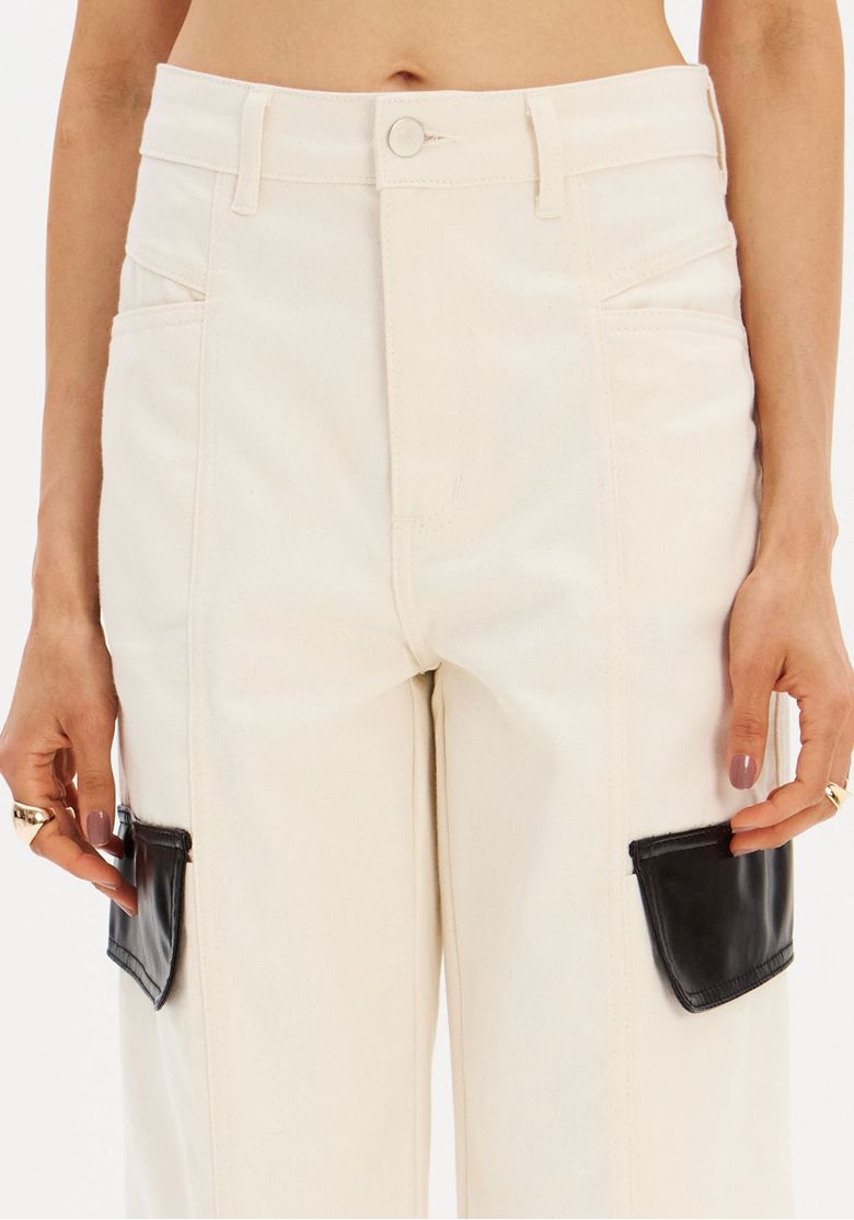 Detalhe frontal da calça de sarja bege com bolsos decorativos em PU e cós com passantes.