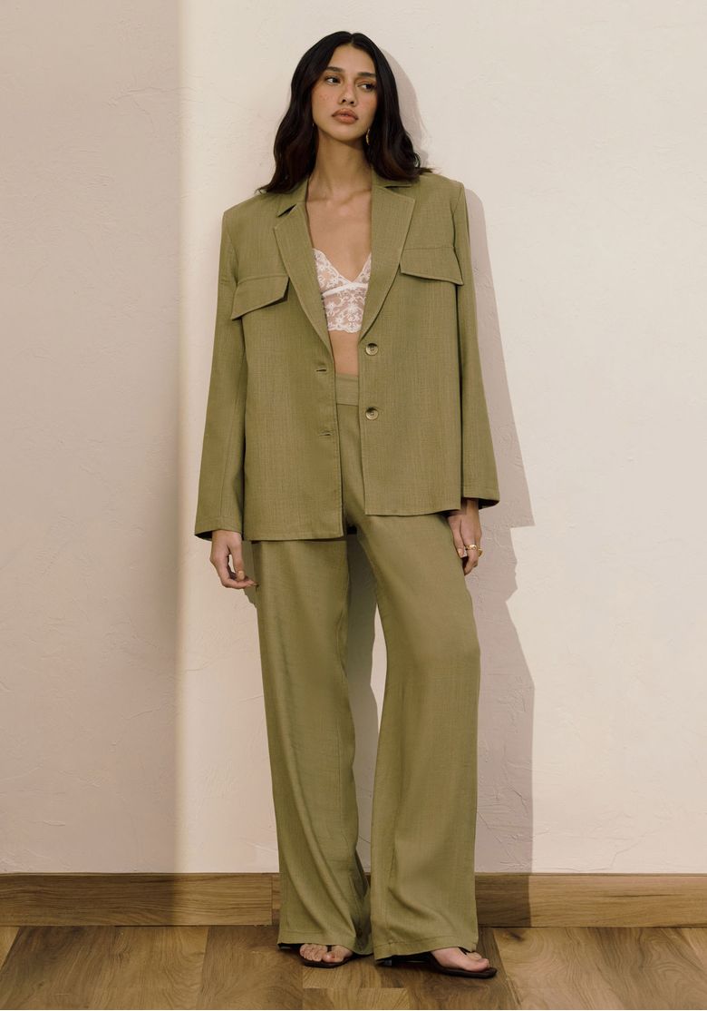 Modelo veste blazer alongado verde em tecido linho-modal, vista frontal com bolsos com aba e dois botões, look completo