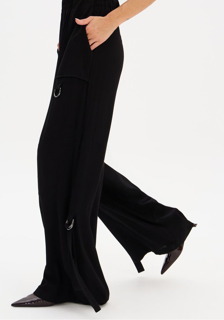 Visão lateral em movimento da calça wide leg preta, destacando tiras decorativas, argola e bolso lateral funcional