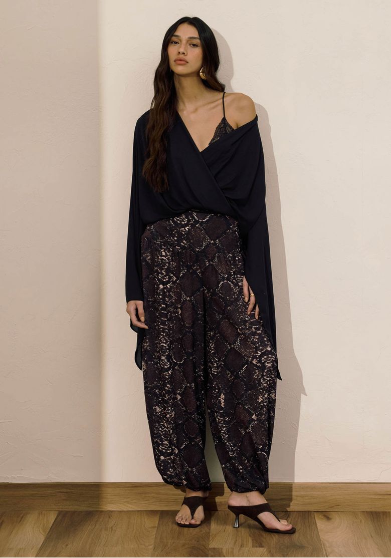 Modelo vestindo uma calça Aladim estampada com estampa croco marrom e cós alto, acompanhada de uma blusa preta drapeada. Look boho fotografado em ambiente interno.