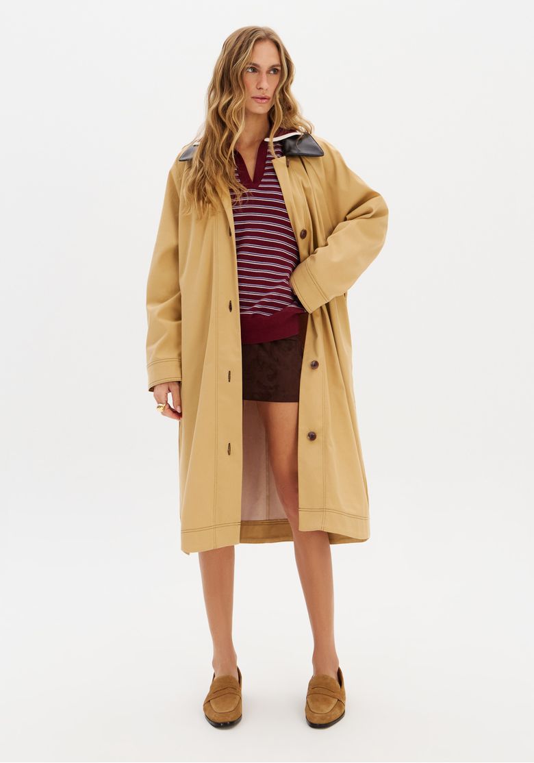 Modelo vestindo trench coat alongado bege com gola preta e faixa, vista frontal com abertura.