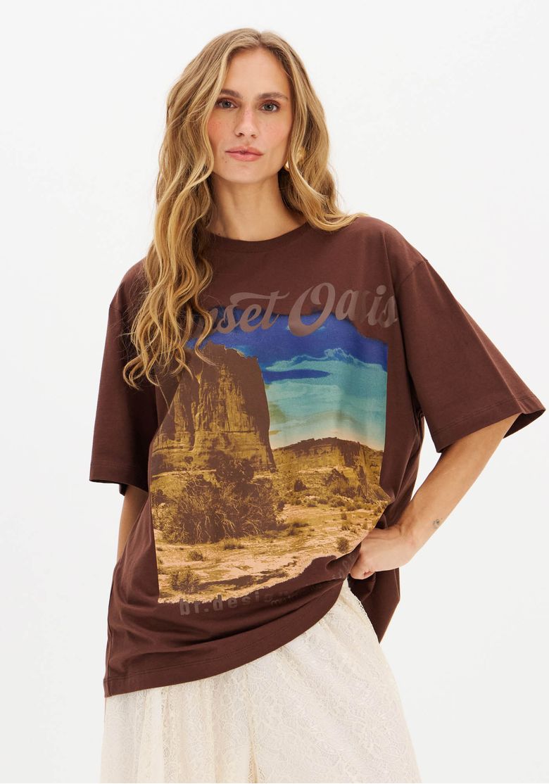 Close frontal de modelo usando camiseta oversized feminina marrom estampada 'Desert Oasis' com destaque para a estampa desértica em tons terrosos e azul.