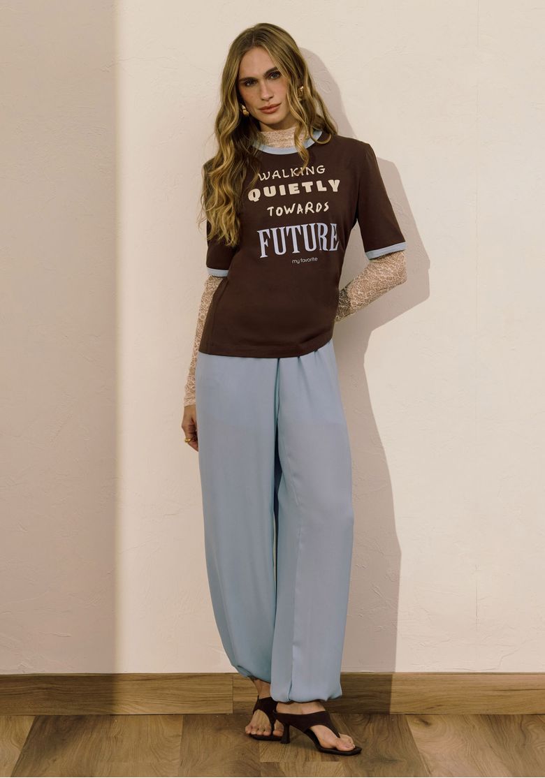 Modelo vestindo camiseta marrom com estampa frontal e calça azul em pose descontraída.