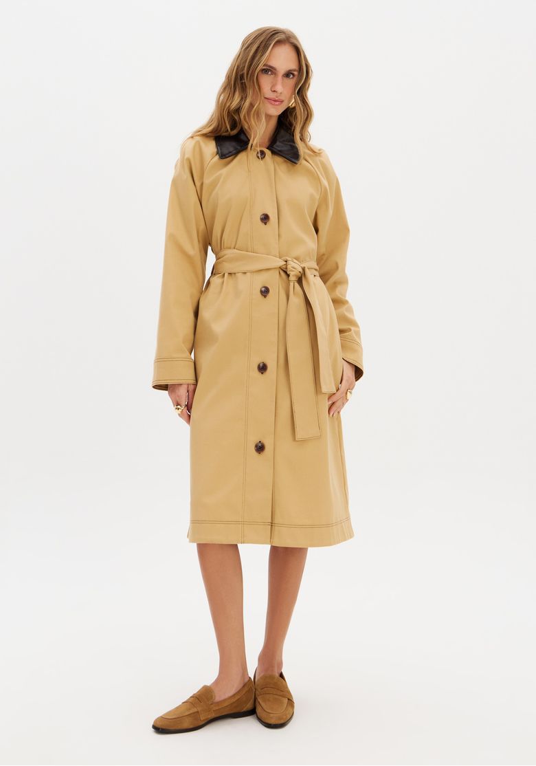 Modelo vestindo trench coat alongado bege com gola preta e faixa, vista frontal com fechamento.