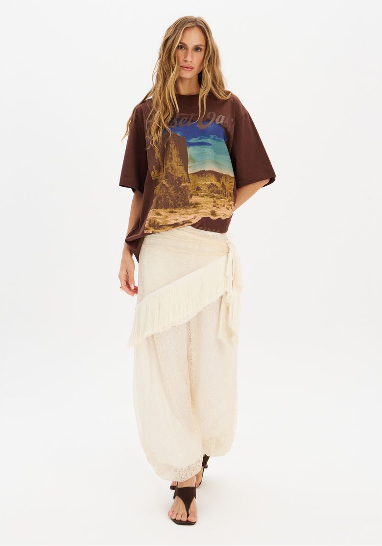 Imagem de corpo inteiro da modelo com camiseta oversized marrom estampada, combinada com saia longa creme e sandálias, visual casual urbano em estúdio.