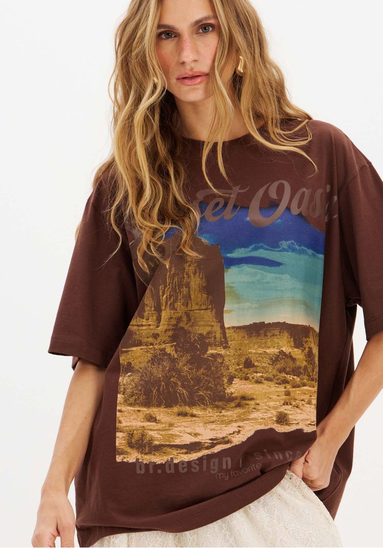 Detalhe de frente da camiseta marrom oversized mostrando a estampa fotográfica paisagem desértica e tipografia, gola redonda e mangas amplas.