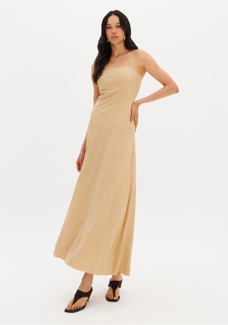 Modelo em fundo branco usando vestido midi sem alça bege, pose três quartos destacando caimento evasê e comprimento midi