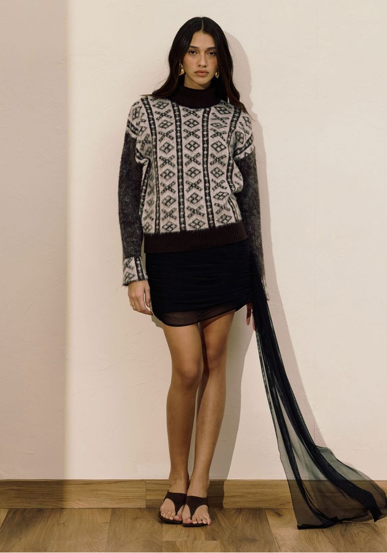 Modelo morena vestindo blusa de tricô gola alta, composê cinza com padronagem geométrica, combinada com saia preta e tecido translúcido longo em ambiente interno.