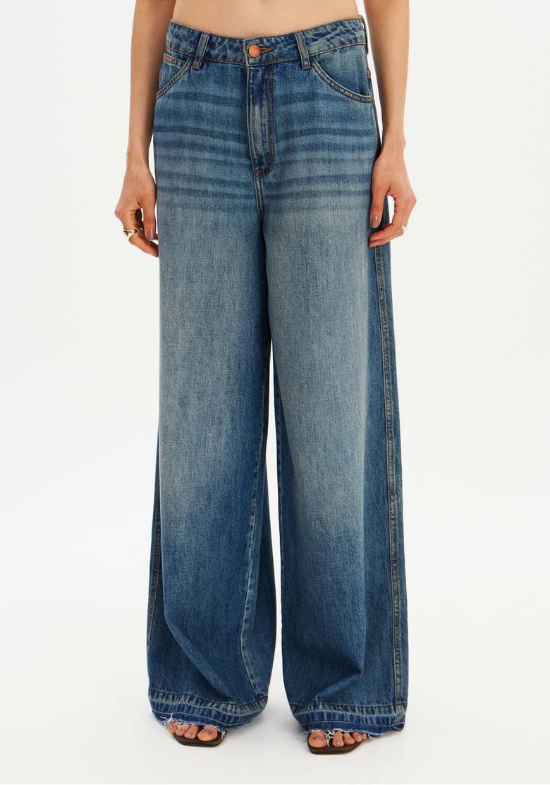 Close frontal da calça jeans relaxed cintura média mostrando cós com passantes, fechamento por zíper e perna ampla com caimento longo