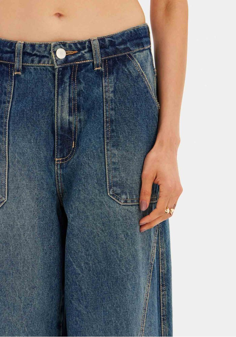 Close-up do cós da calça jeans barrel leg azul com passantes e botão.