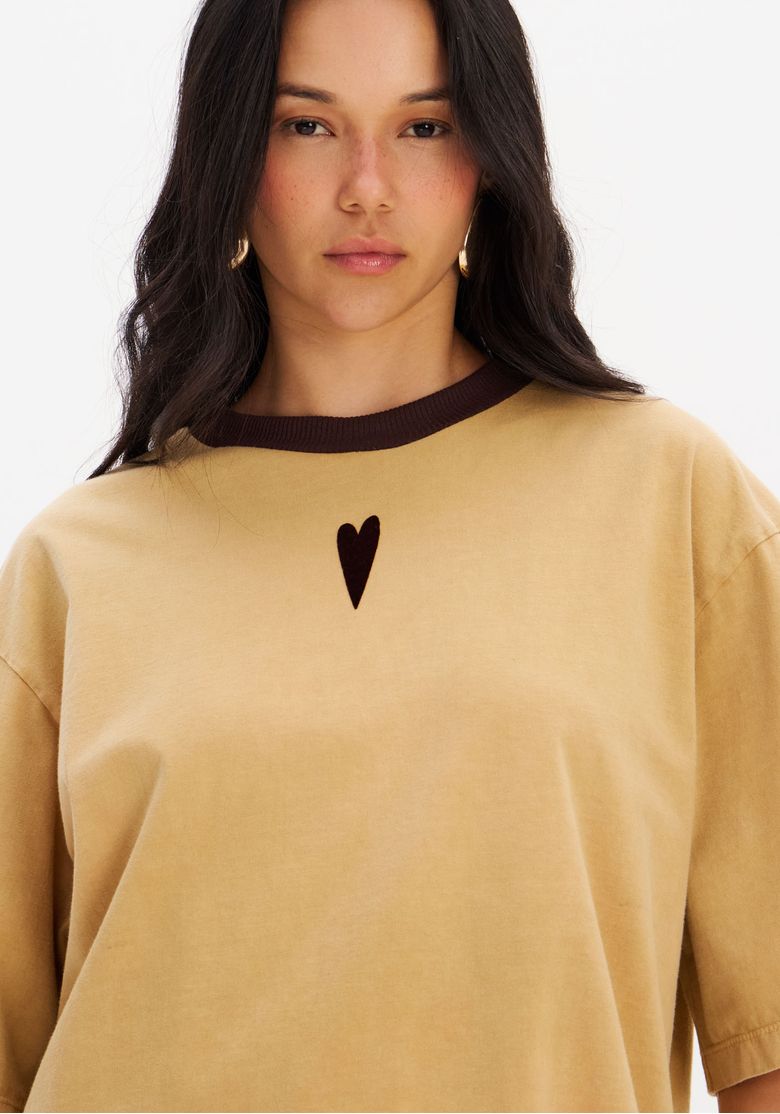 Visão média da camiseta oversized bege com detalhe da estampa de coração central e gola redonda com rib marrom, evidenciando o caimento amplo.