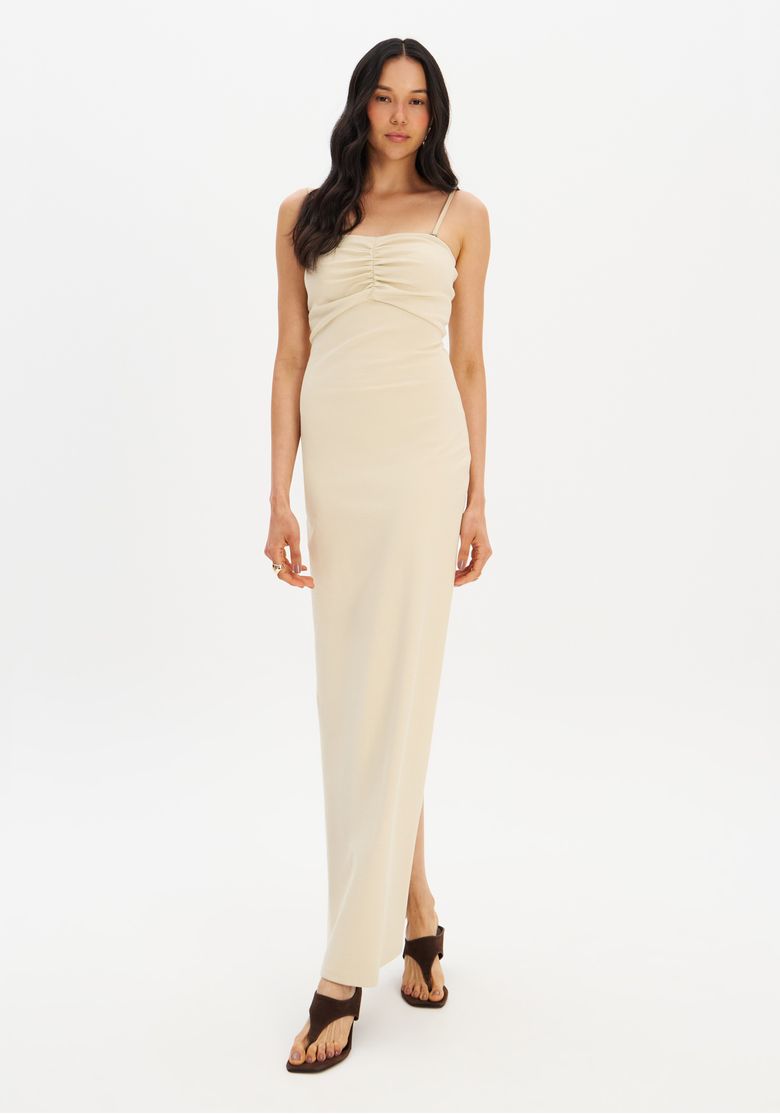 Modelo vestindo o vestido super midi de malha bege em uma vista frontal corpo inteiro mostrando o caimento justo, alças finas ajustáveis e uma fenda discreta na lateral das costas.