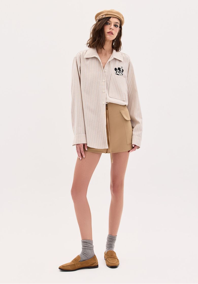 Modelo vestindo camisa feminina Disney com listras bege e bordado do Mickey, combinada com shorts bege.
