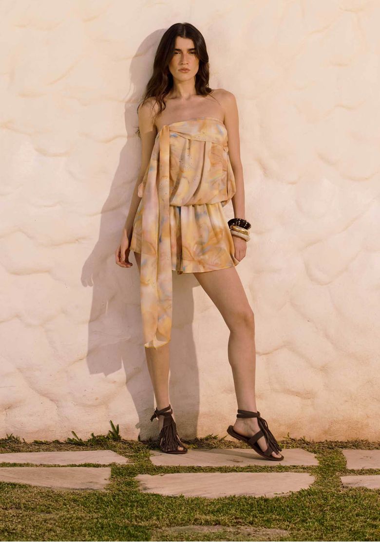 Modelo com macacão curto feminino sem alças amarelo estampado em proposta sunset, com caimento fluido e detalhe lateral longo