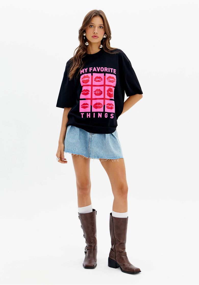 Modelo vestindo saia jeans curta com pregas e camisa preta estampada, combinada com botas marrons.