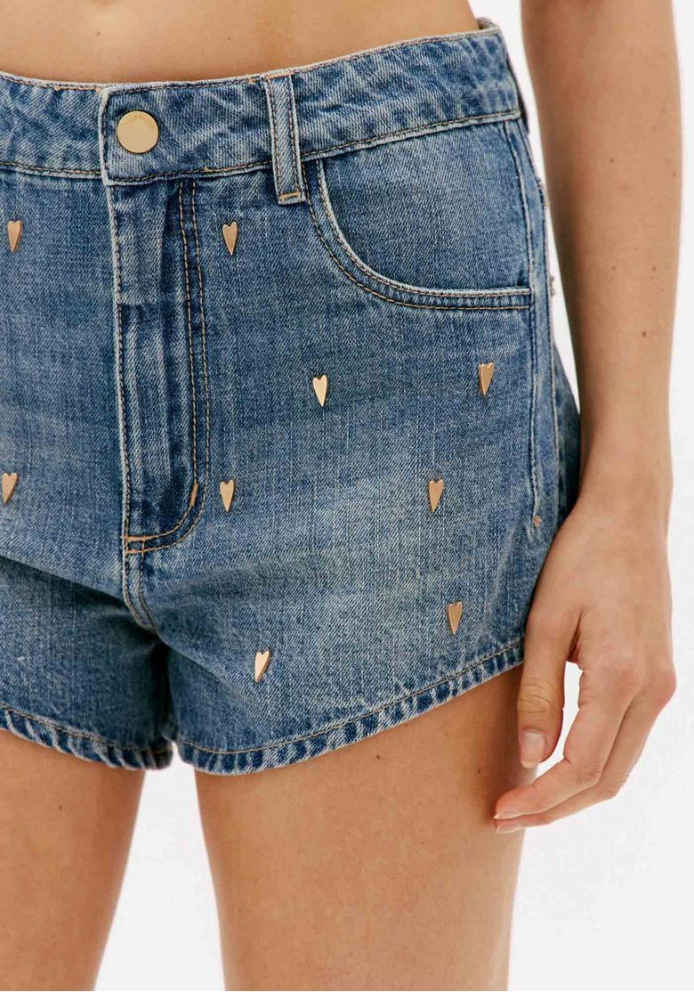 Close no short jeans azul com aplicações de coração, cós com passantes, bolso frontal e fechamento por botão