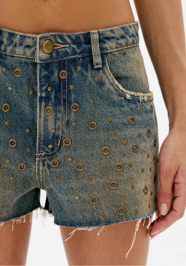 Close no short jeans azul feminino com cintura baixa, botão frontal, passantes, aplicações metálicas e barra desfiada