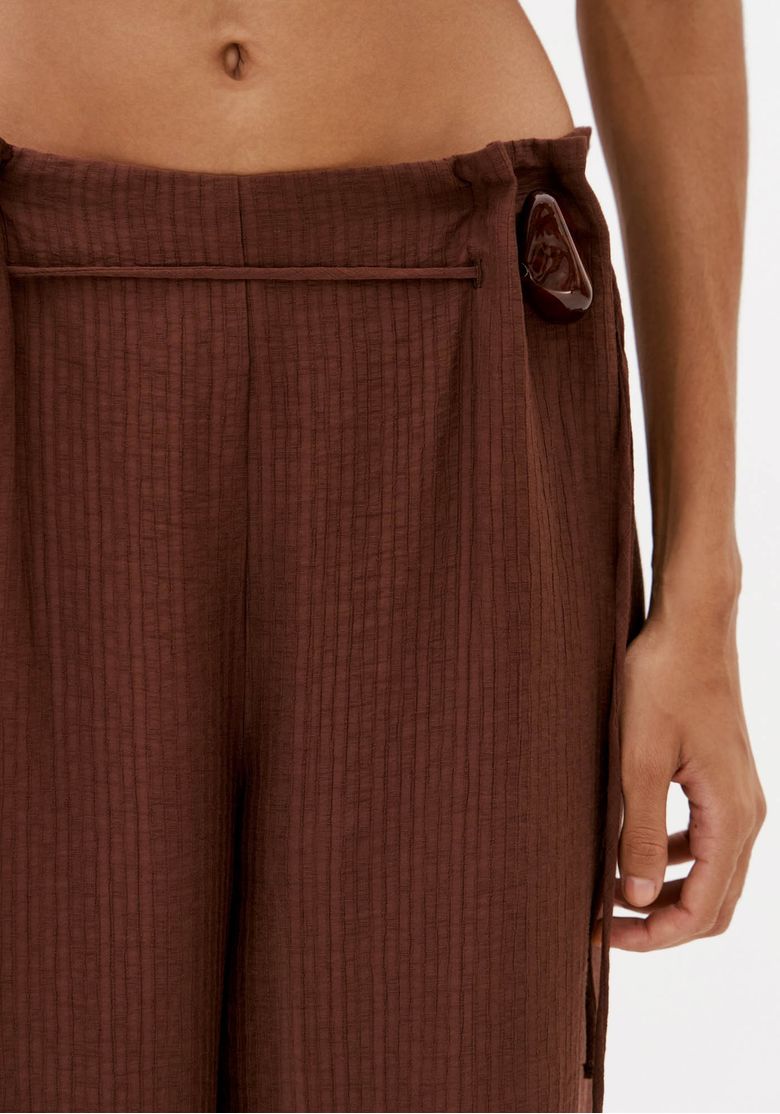 Close da cintura da calça marrom com pregas frontais, cordão lateral e detalhe de aviamento decorativo