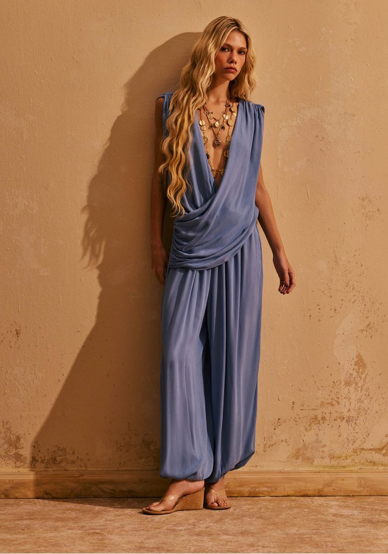 Modelo usando macacão longo aladim azul claro com decote drapeado profundo e sandália rasteira, em look elegante para sunset